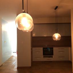TOM BIN-REMONTY MIESZKAN POD KLUCZ - Nowoczesna kuchnia z drewnianą podłogą, jasnymi szafkami i okładziną, oświetlona przez dwie wiszące lampy z okrągłymi, szklanymi kloszami.