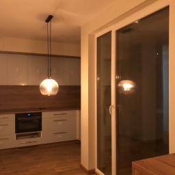 TOM BIN-REMONTY MIESZKAN POD KLUCZ - Nowoczesna kuchnia z jasnymi szafkami, drewnianym blatem, podłogą z paneli i szklaną lampą wiszącą, widok na zewnątrz przez duże okno balkonowe.