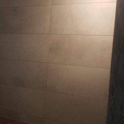 TOM BIN-REMONTY MIESZKAN POD KLUCZ - Ściana wyłożona beżowymi, podłużnymi płytkami ceramicznymi, z widocznym fragmentem drewnopodobnej podłogi i wystającym kablem elektrycznym u góry.