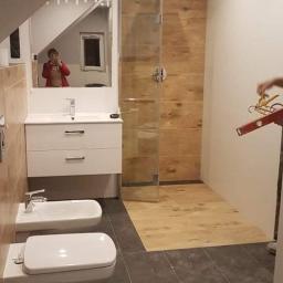 TOM BIN-REMONTY MIESZKAN POD KLUCZ - Łazienka w trakcie remontu na poddaszu: kabina prysznicowa z drewnopodobnymi płytkami, biała umywalka z szafką, bidet i toaleta na ciemnych płytkach. W lustrze odbija się osoba robiąca zdjęcie.