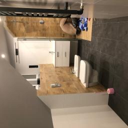 TOM BIN-REMONTY MIESZKAN POD KLUCZ - Wnętrze łazienki w trakcie prac wykończeniowych: widoczna umywalka z szafką, toaleta, bidet, grzejnik drabinkowy, płytki imitujące drewno na podłodze i pod prysznicem oraz ciemne płytki na ścianach...