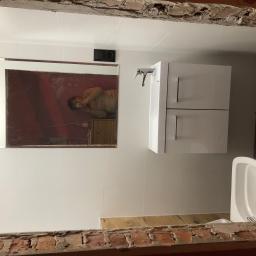 TOM BIN-REMONTY MIESZKAN POD KLUCZ - Nowoczesna umywalka z szafką w łazience w trakcie remontu, widoczne białe płytki i surowy mur pod sufitem, odbicie fotografa w lustrze.