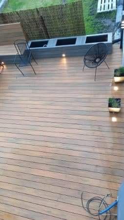 Drewniany taras z desek kompozytowych, oświetlenie punktowe, metalowe krzesła i donice z zielenią, widok z góry, fragment węża ogrodowego w rogu.