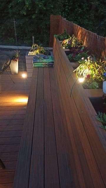 Nowoczesny taras z desek kompozytowych, podświetlony punktowymi lampami, z wbudowanymi rabatami kwiatowymi i ozdobnym bambusowym płotem w tle.