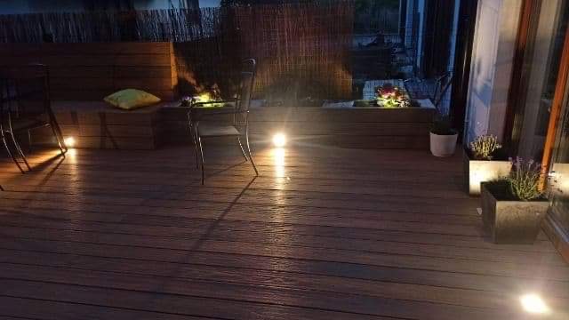 Ciemna, drewniana podłoga tarasu z podświetleniem punktowym, meblami ogrodowymi i roślinami w donicach, oświetlona ciepłym światłem.