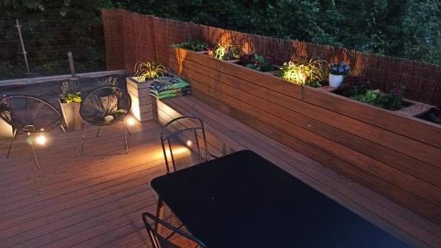 Drewniany taras z podwyższonymi rabatami, oświetleniem punktowym, dwoma ażurowymi fotelami i czarnym stołem, oświetlony wieczorową porą.