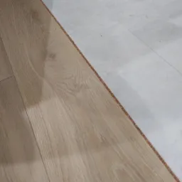 Fragment podłogi ukazujący połączenie paneli podłogowych z jasnymi, kwadratowymi płytkami ceramicznymi; widoczna struktura drewna i delikatna faktura płytek.