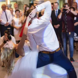 Para młoda w tańcu podczas wesela, panna młoda w sukni z gorsetem, pan młody w białej koszuli i granatowych spodniach, w tle goście i fotografowie.