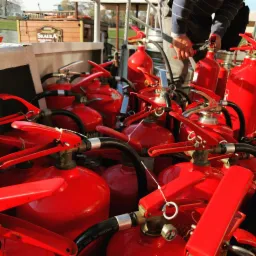 Grupowe zdjęcie wielu czerwonych gaśnic proszkowych, prawdopodobnie podczas kontroli lub przygotowania do szkolenia, z widoczną ręką osoby sprawdzającej jedną z nich.