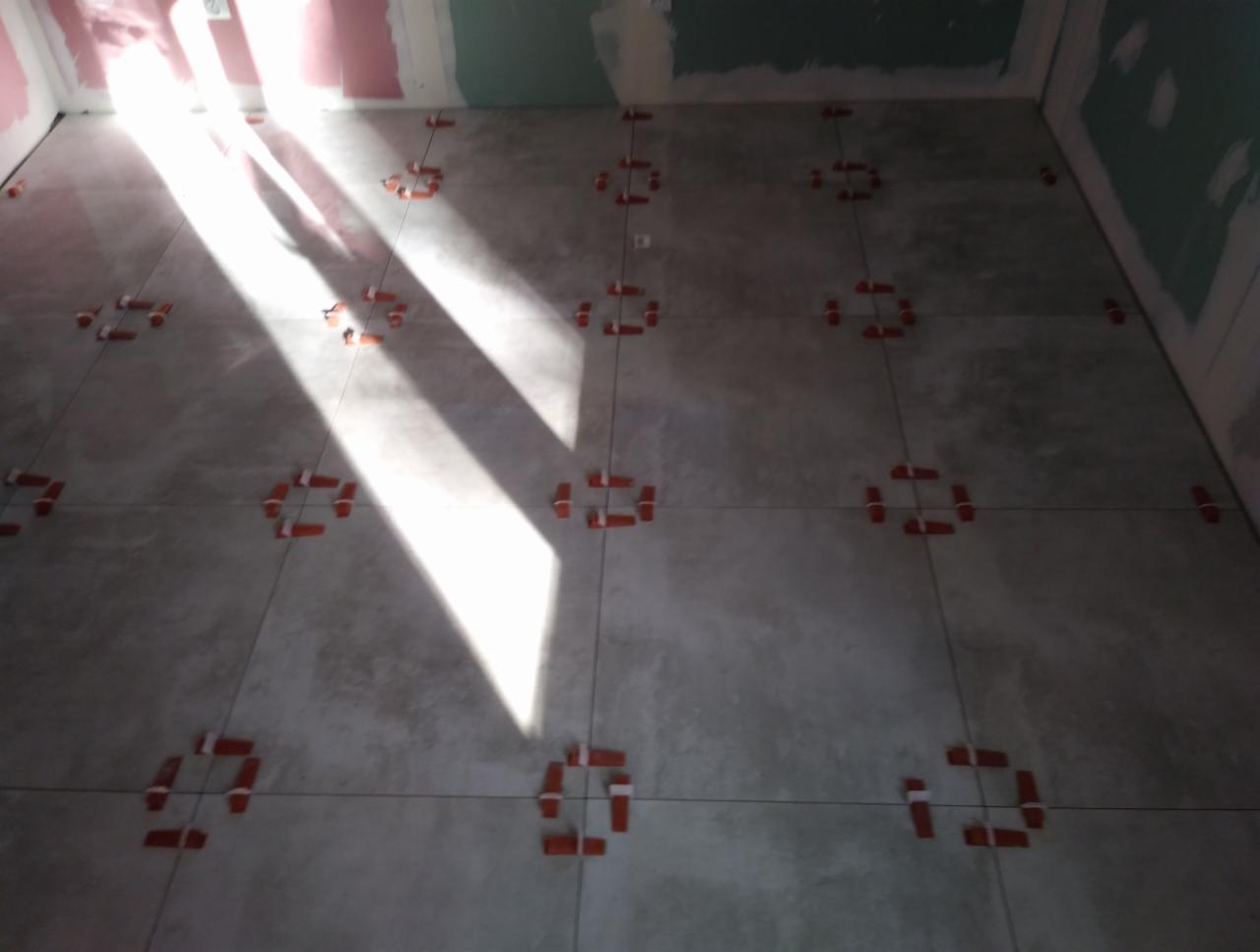 Układanie szarych płytek podłogowych z użyciem pomarańczowych krzyżyków dystansowych, oświetlone mocnym światłem słonecznym padającym z okna.