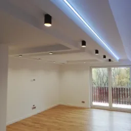 Nowoczesne wnętrze z drewnianą podłogą, białymi ścianami i sufitem podwieszanym z oświetleniem LED oraz punktowymi lampami w kolorze czarnym, widok na balkon z drewnianą balustradą i drzewa za oknem.