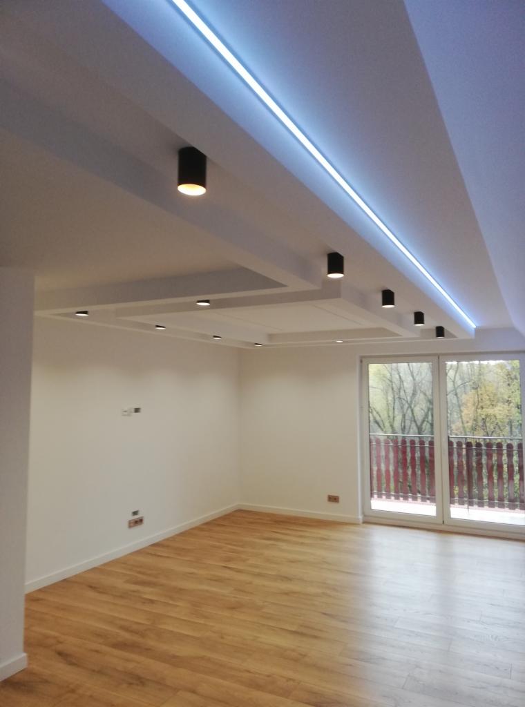 Nowoczesne wnętrze z drewnianą podłogą, białymi ścianami i sufitem podwieszanym z oświetleniem LED oraz punktowymi lampami w kolorze czarnym, widok na balkon z drewnianą balustradą i drzewa za oknem.