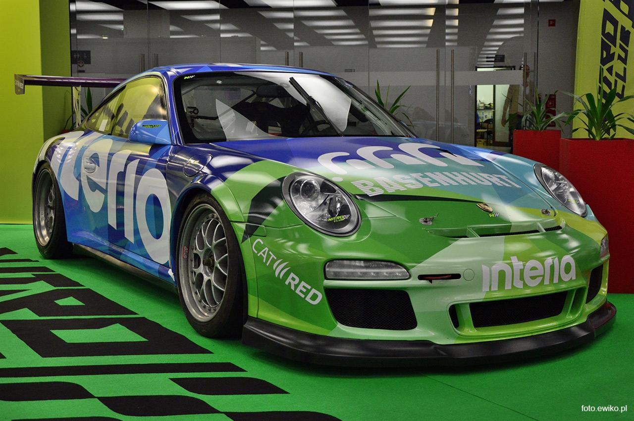 Porsche 911 GT3 Cup z barwnym oklejeniem w stylu wyścigowym, zaparkowane na zielonej podłodze z czarnymi wzorami, widoczne logo 'Interia' i 'CAT RED'.