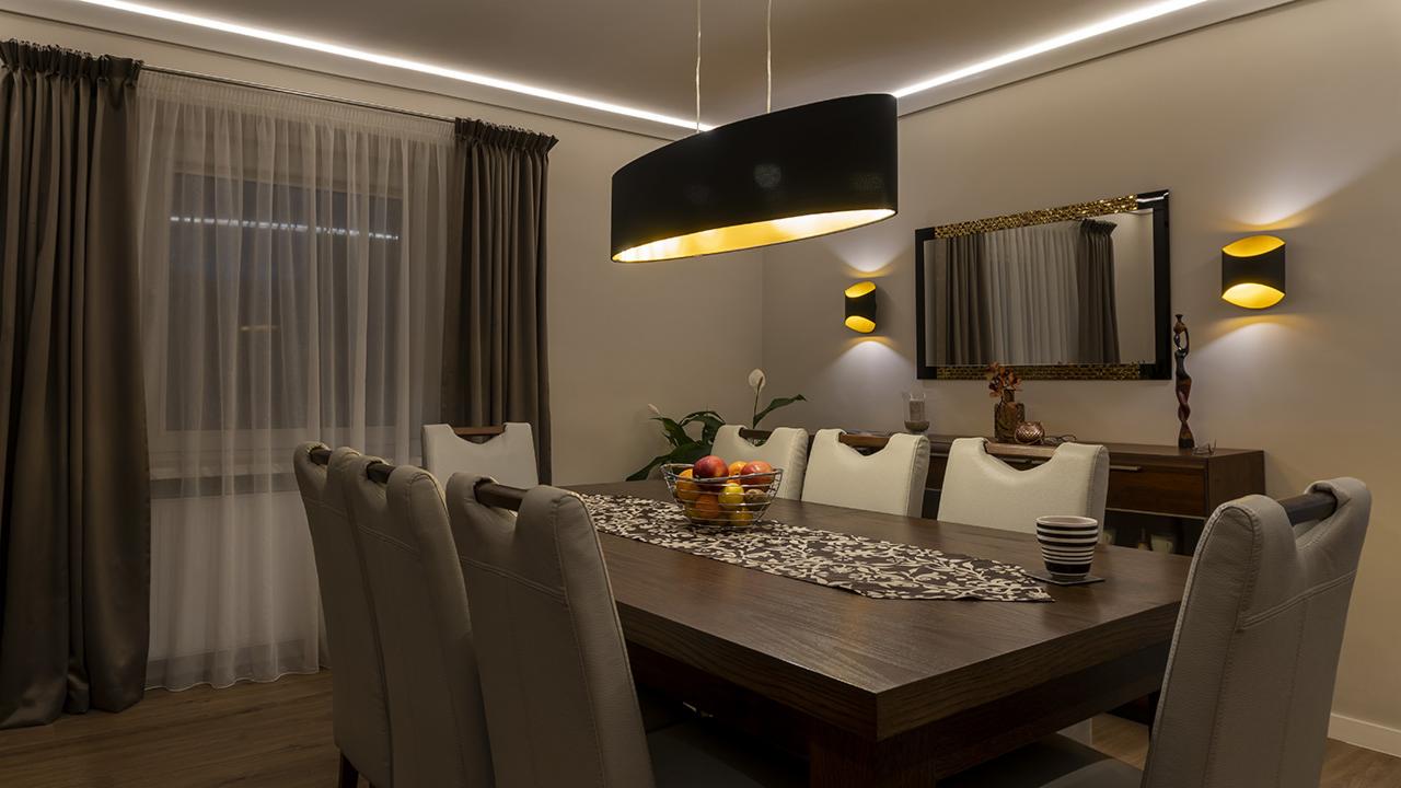 Jadalnia z ciemnym drewnianym stołem, nakrytym bieżnikiem w kwiaty i misą owoców, oświetlona wiszącą czarną lampą z wnętrzem w kolorze złota i kinkietami.