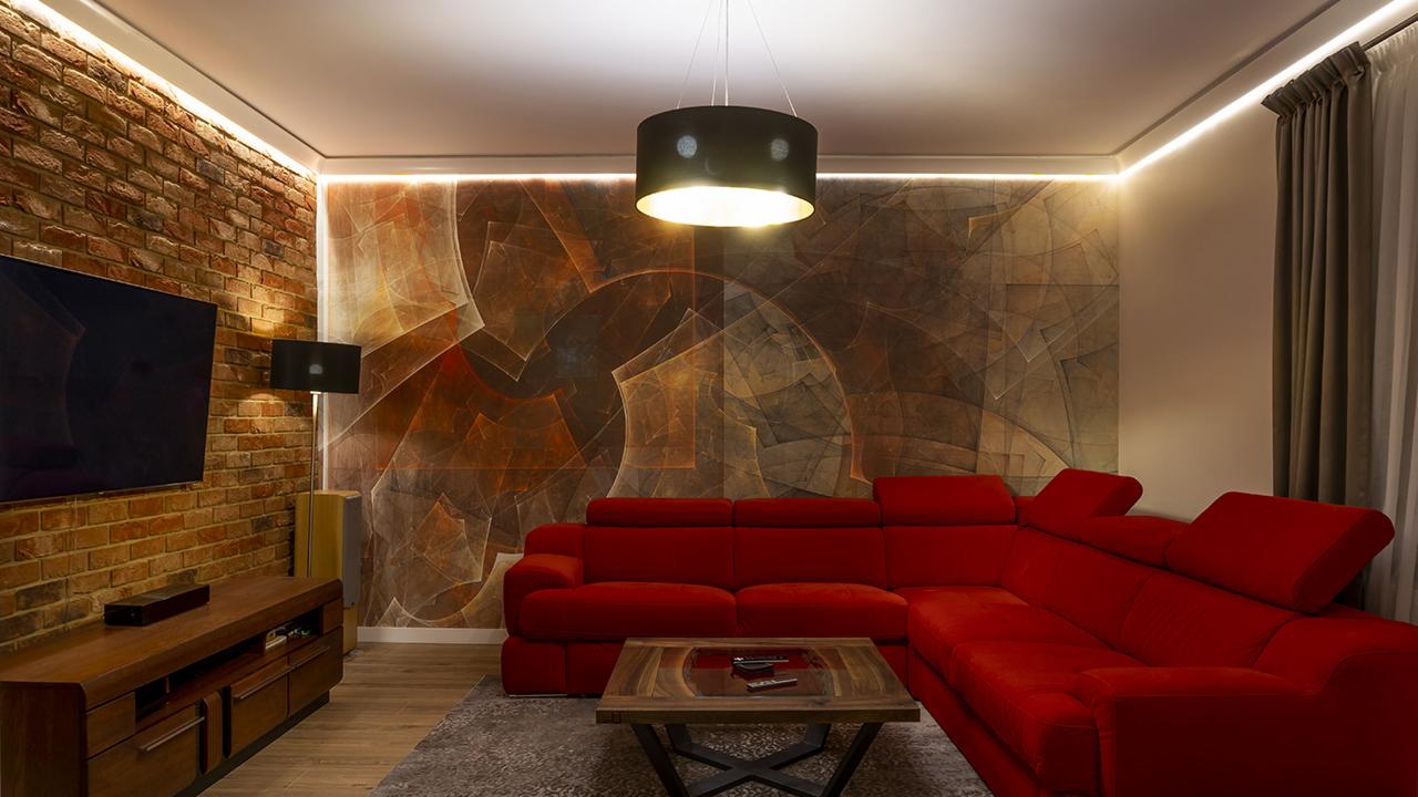 Nowoczesny salon z czerwoną sofą narożną, ceglaną ścianą, abstrakcyjną tapetą i designerskim oświetleniem.