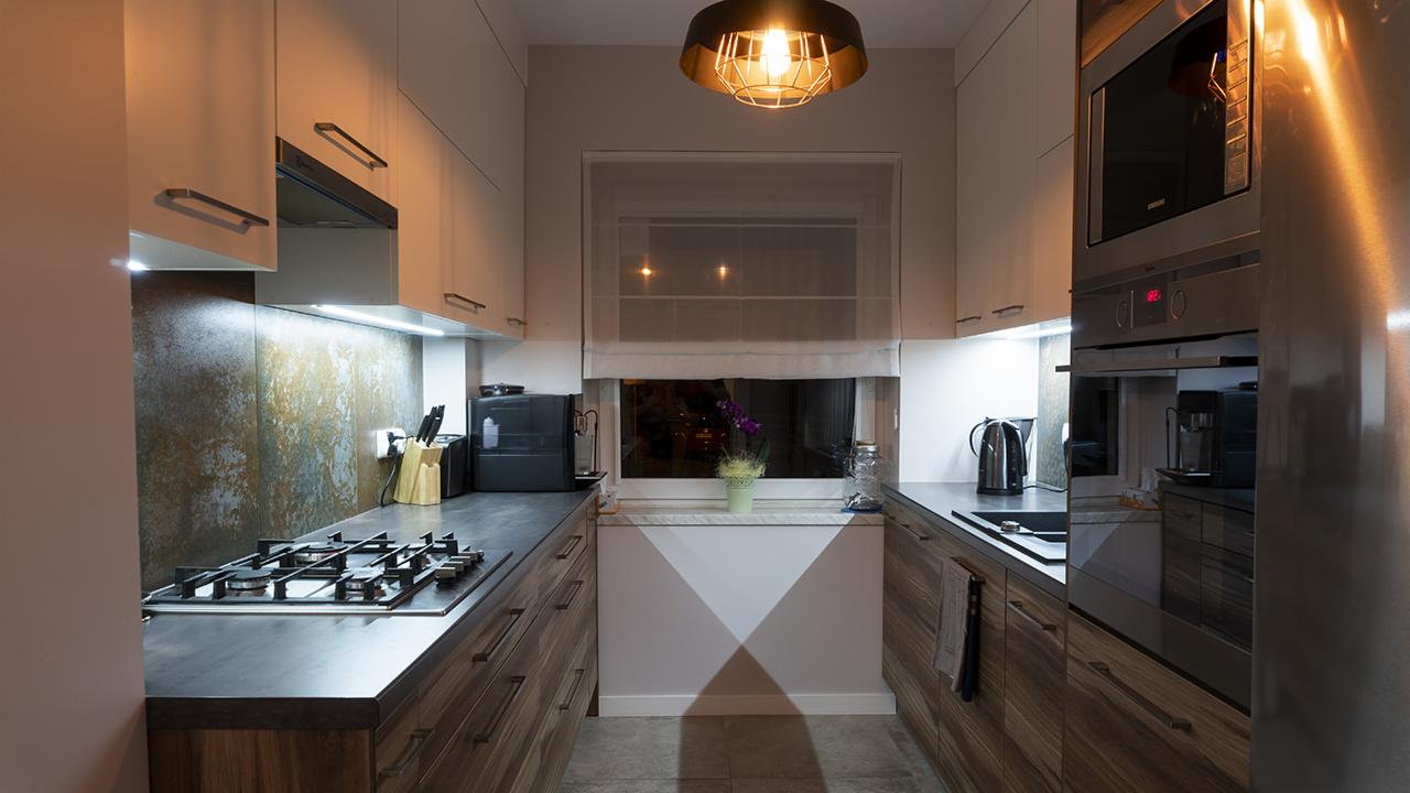 Wnętrze nowoczesnej kuchni z drewnianymi szafkami i szarym blatem, oświetlone lampą wiszącą z metalowym kloszem i dodatkowym oświetleniem LED pod szafkami.