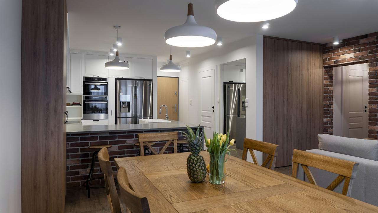 Nowoczesna kuchnia z wyspą wykończoną cegłą, drewnianym stołem z ananasem i tulipanami w wazonie, oświetlona wiszącymi lampami w kształcie kopuł.