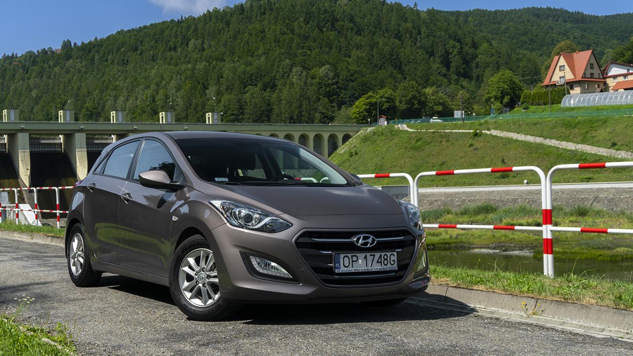 Brązowy Hyundai i30 na tle zapory wodnej i zalesionego wzgórza, tablica rejestracyjna OP 1748G.