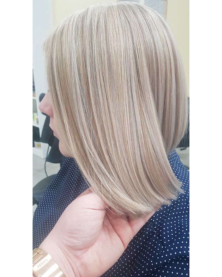 Blond włosy z delikatnymi pasemkami, świeżo po strzyżeniu na boba, widok z profilu na tle granatowej bluzki w białe kropki i ręki z zegarkiem.