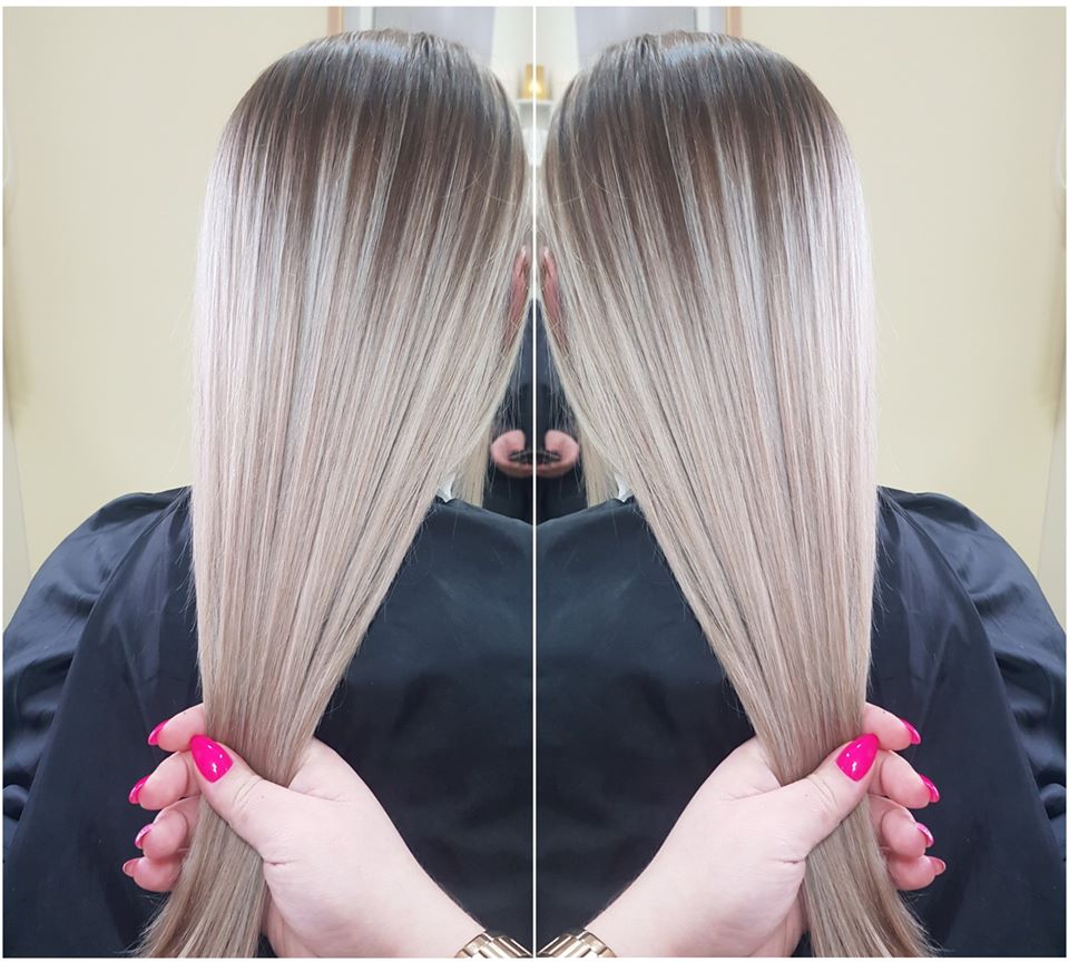 Długie, proste włosy z ombre od ciemnego brązu u nasady do jasnego blondu na końcach, trzymane w dłoniach z jaskraworóżowym manicure, odbicie w lustrze w tle.