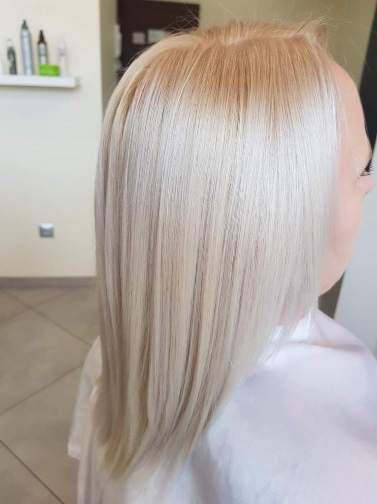 Proste, platynowe blond włosy z delikatnym odrostem w odcieniu ciepłego blondu, widoczne z profilu na tle salonu.