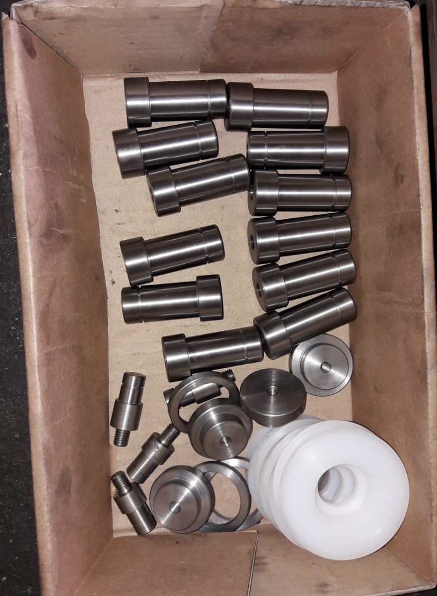 Kartonowe pudełko wypełnione metalowymi i plastikowymi elementami o różnych kształtach i rozmiarach, w tym cylindrycznymi wałkami, pierścieniami i dyskami. Elementy są ułożone chaotycznie,...