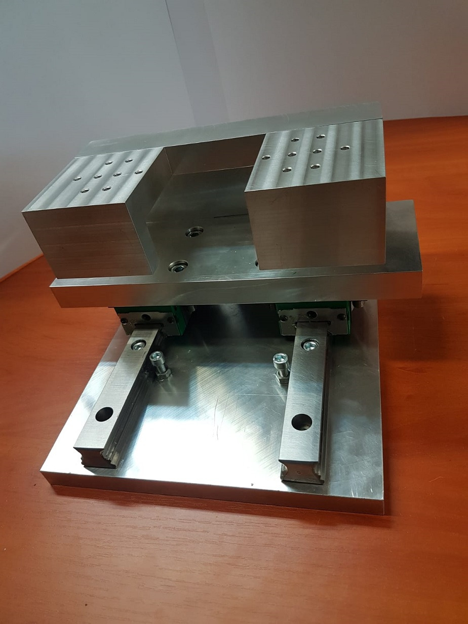 Metalowa konstrukcja z otworami i śrubami, prawdopodobnie element maszyny, na drewnianym stole, oświetlona z góry.