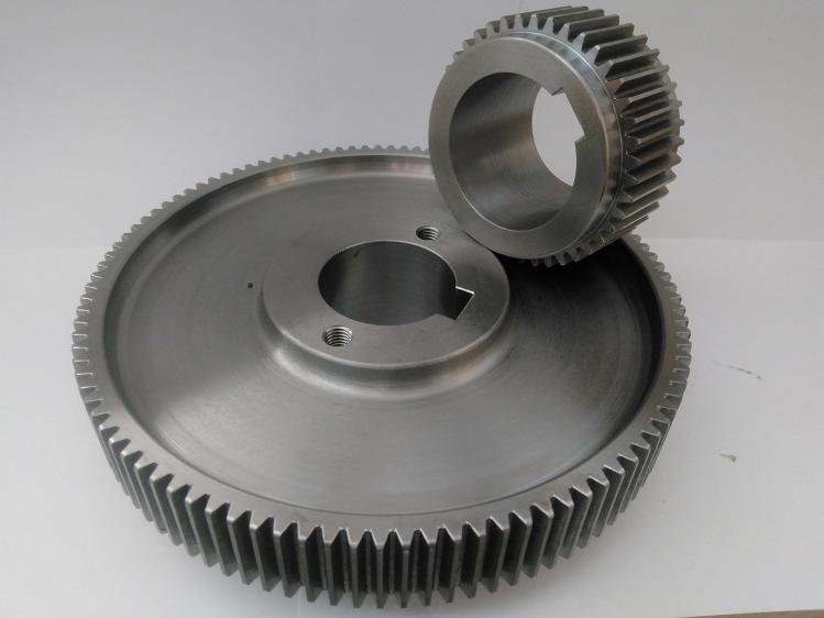 Dwie metalowe zębatki o różnej wielkości, większa z trzema otworami gwintowanymi wokół centralnego otworu, mniejsza ustawiona pionowo za większą, na neutralnym tle.