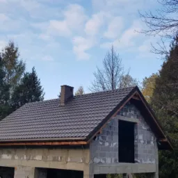 Dom w trakcie budowy z brązowym dachem z dachówki, widoczne bloczki betonowe i komin, otoczony drzewami na tle błękitnego nieba z chmurami.