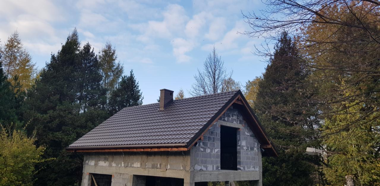 Dom w trakcie budowy z brązowym dachem z dachówki, widoczne bloczki betonowe i komin, otoczony drzewami na tle błękitnego nieba z chmurami.