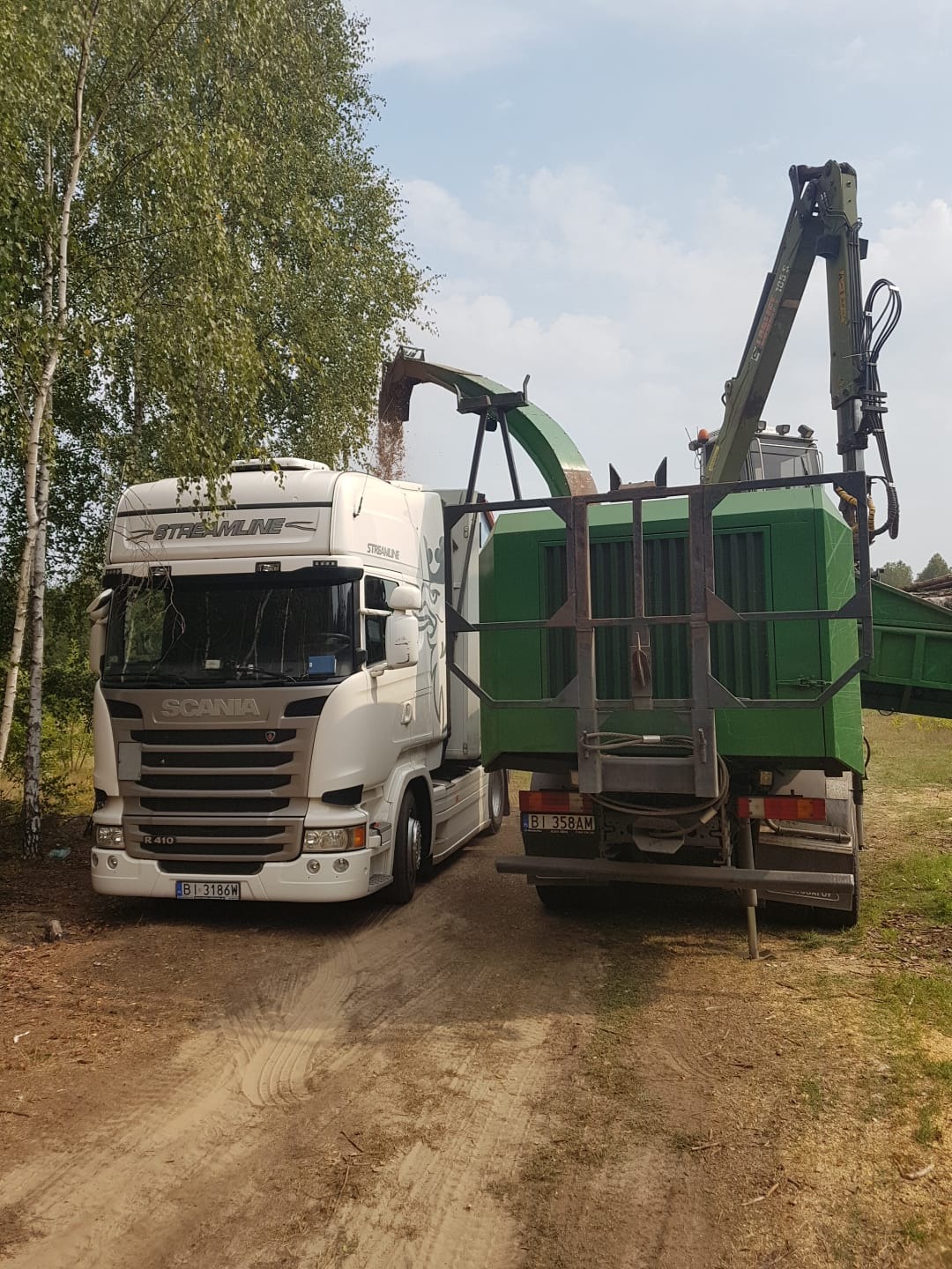 Biała ciężarówka Scania Streamline R410 z podpiętą zieloną rębarką do gałęzi podczas załadunku zrębków na leśnej drodze.