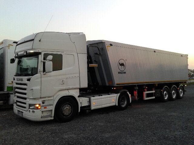 Biała ciężarówka Scania z naczepą marki Wielton, widoczny symbol wielbłąda na naczepie, zaparkowana na żwirowym placu o zmierzchu.