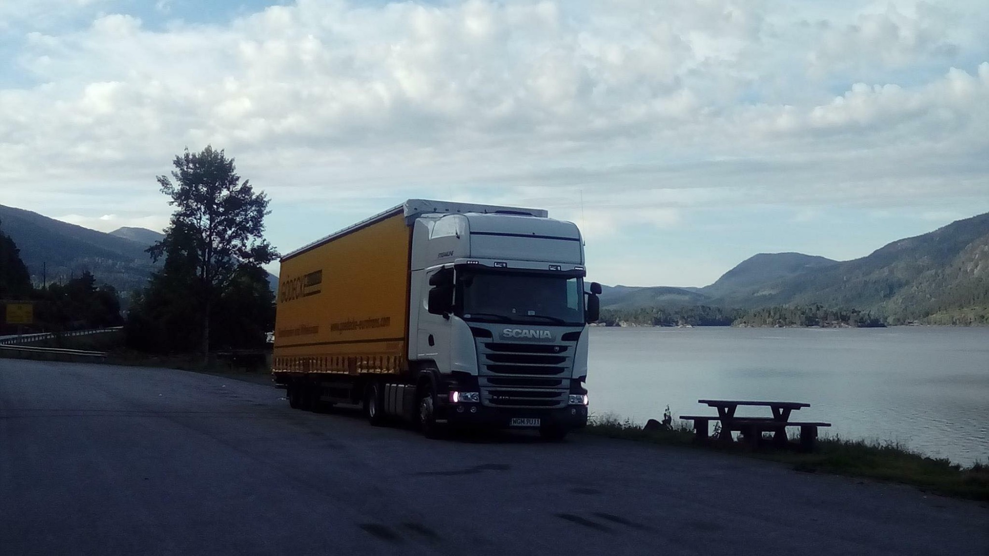 Żółta ciężarówka Scania z naczepą na tle jeziora i gór, widok z boku, pochmurne niebo.