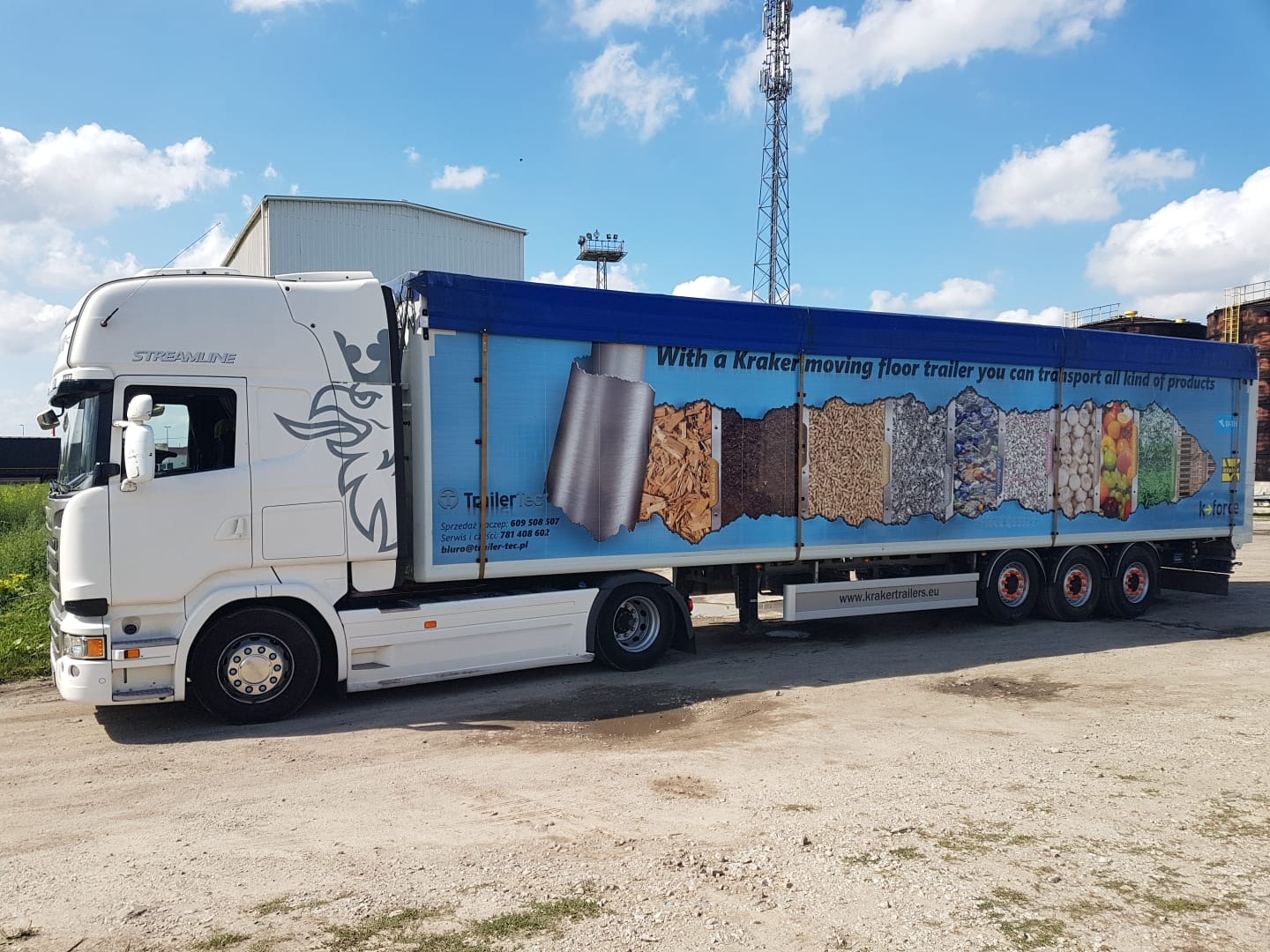 Biała ciężarówka Scania Streamline z niebieską naczepą Kraker z grafiką przedstawiającą różne rodzaje ładunków, zaparkowana na placu, widoczna wieża telekomunikacyjna w tle.