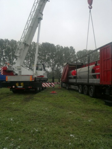 Żuraw przenosi element konstrukcyjny do czerwonej naczepy ciężarówki na zewnątrz, w tle widoczne drzewa i pochmurne niebo.