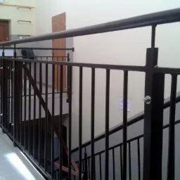 Ciemnobrązowa, metalowa balustrada schodowa z pionowymi szczeblami, mocowana na śruby, widok z boku, ściana w tle w kolorze złamanej bieli.