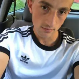 Młody mężczyzna w koszulce Adidas z trzema paskami siedzący w samochodzie, z widocznym pasem bezpieczeństwa i zagłówkiem.