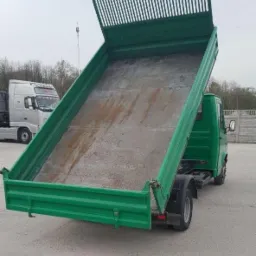 Zielona wywrotka z podniesioną skrzynią ładunkową, widoczne ślady użytkowania wewnątrz, na betonowym placu, z białym ciągnikiem siodłowym w tle.