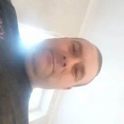 Mężczyzna w czarnej koszulce z logo GDL, na tle białej ściany. Ujęcie z dołu, widoczna część sufitu i okno. Ściana wydaje się być świeżo pomalowana.