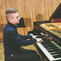 Młody mężczyzna w ciemnej koszuli gra na czarnym fortepianie z otwartą klapą, widoczne struny i mechanizm instrumentu, drewniane panele na ścianie w tle.