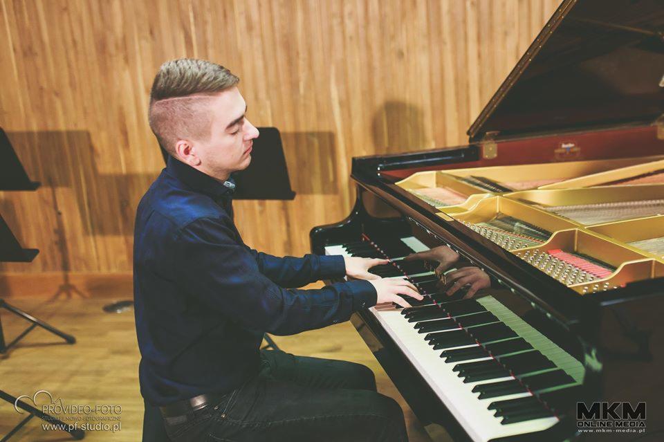 Młody mężczyzna w ciemnej koszuli gra na czarnym fortepianie z otwartą klapą, widoczne struny i mechanizm instrumentu, drewniane panele na ścianie w tle.