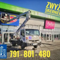 Pracownicy na podnośniku koszowym montują lub czyszczą duży szyld reklamowy na fasadzie sklepu, widoczny pojazd z numerem telefonu kontaktowym.