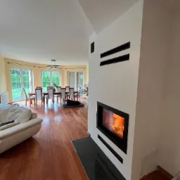 Nowoczesny wkład kominkowy z czarną ramą, wbudowany w białą ścianę z poziomymi, czarnymi elementami dekoracyjnymi, widoczne płomienie, w tle salon z drewnianą podłogą, stołem i krzesłami.