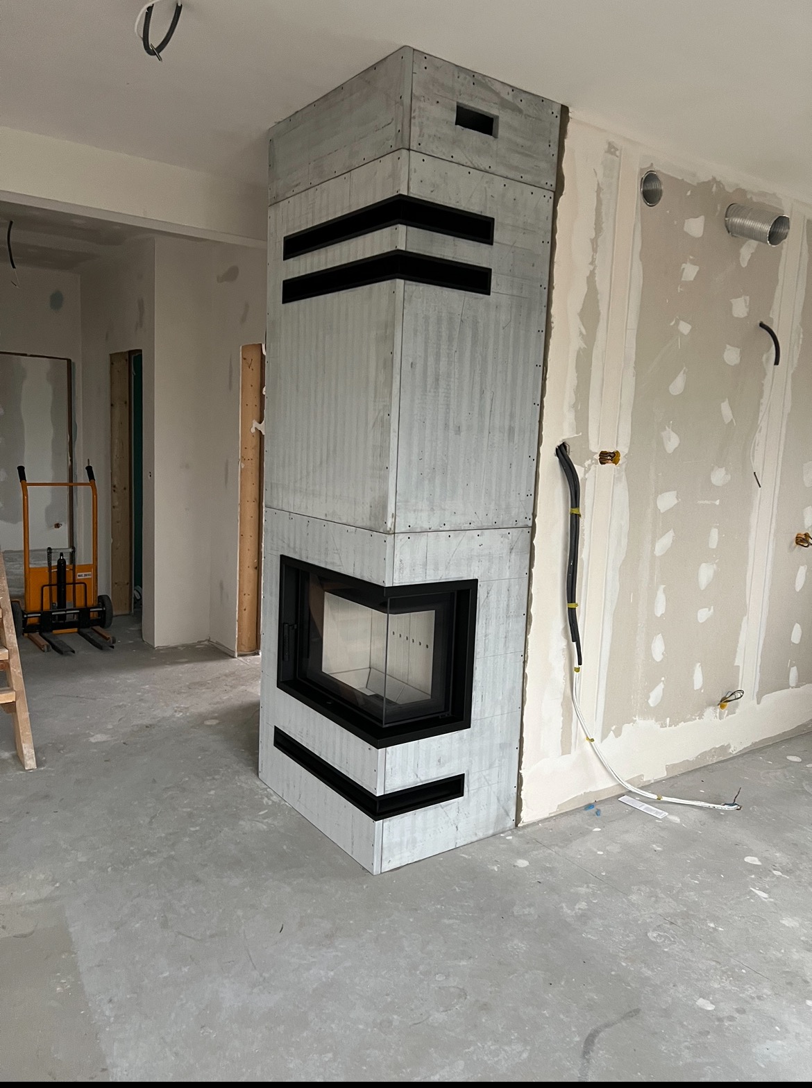 Narożny, betonowy kominek z czarnymi, poziomymi wstawkami w surowym wnętrzu w trakcie wykończenia, widoczne kable i narzędzia.