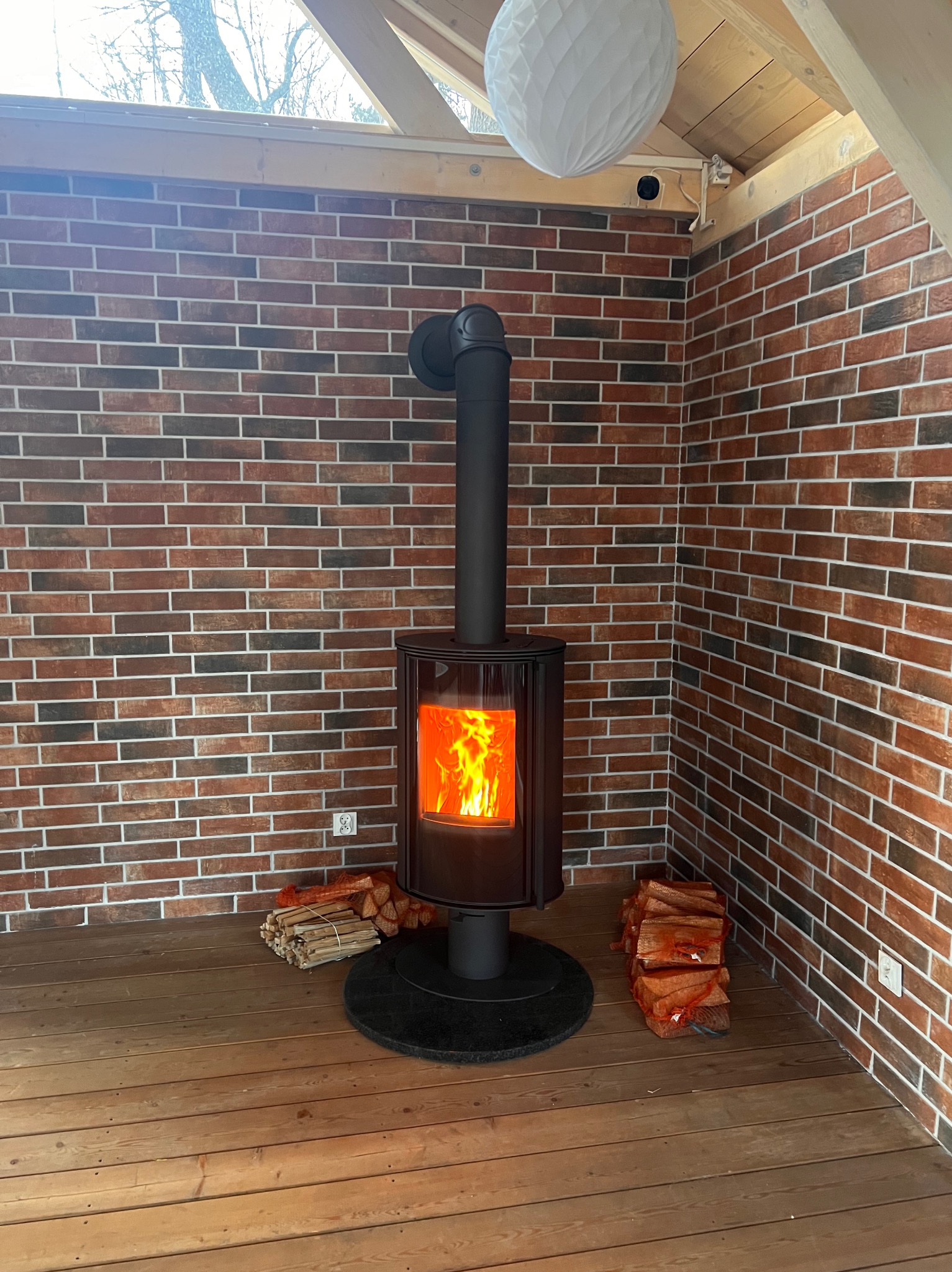 Nowoczesny piec wolnostojący z palącym się ogniem, umieszczony w rogu pomieszczenia wyłożonego cegłą, z zapasem drewna obok.