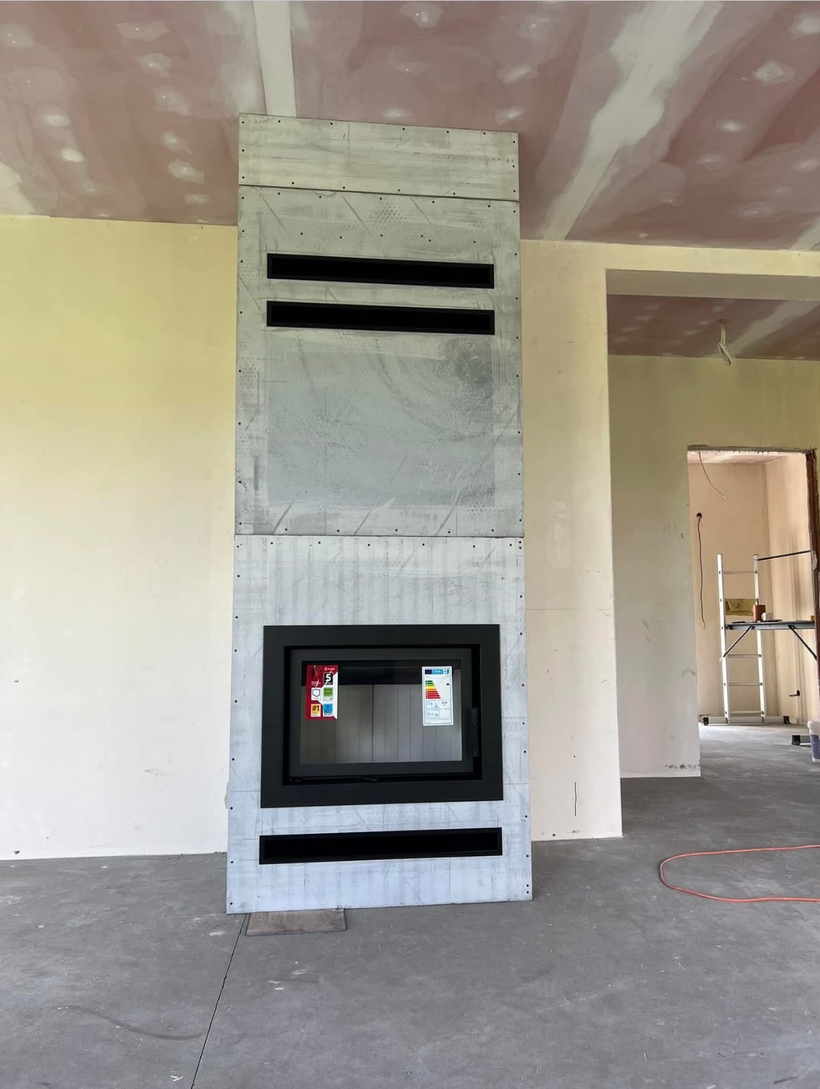 Surowy, wysoki obudowany wkład kominkowy z czarną ramą, w trakcie montażu, widoczne naklejki na szybie, sufit i ściany w stanie deweloperskim.