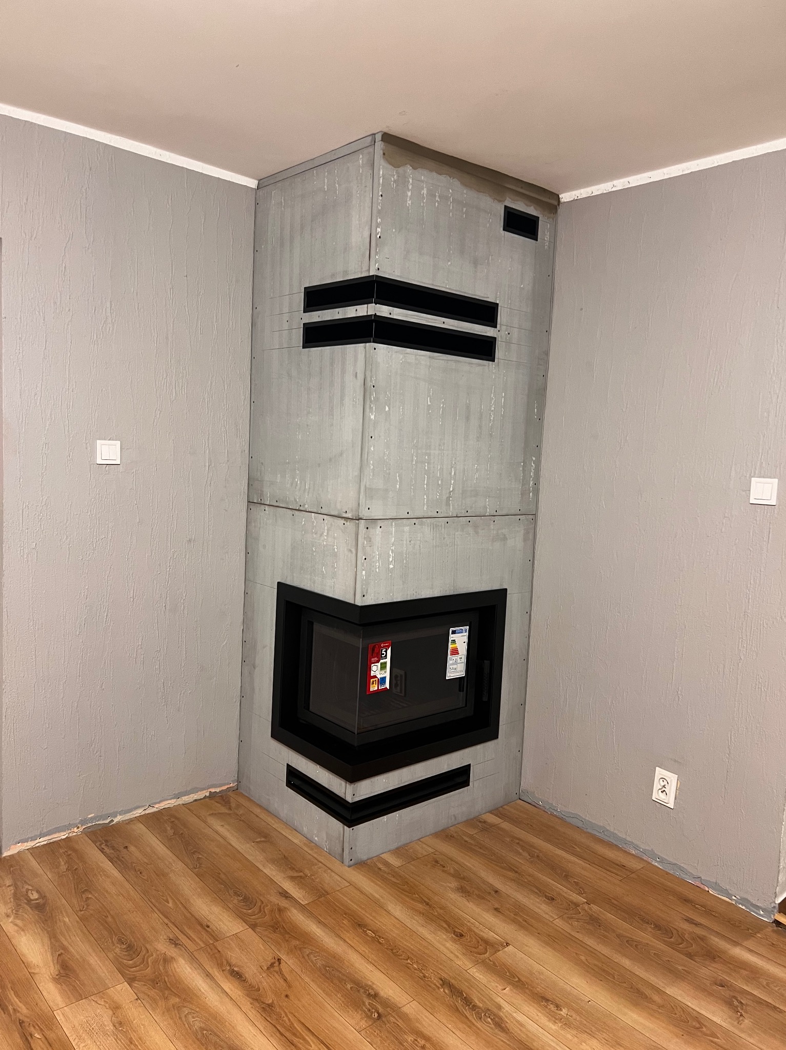 Narożny kominek z szarą obudową z betonu architektonicznego, czarny wkład kominkowy z szybą, widoczne naklejki informacyjne na szybie, szare ściany z fakturą, brązowe panele podłogowe.