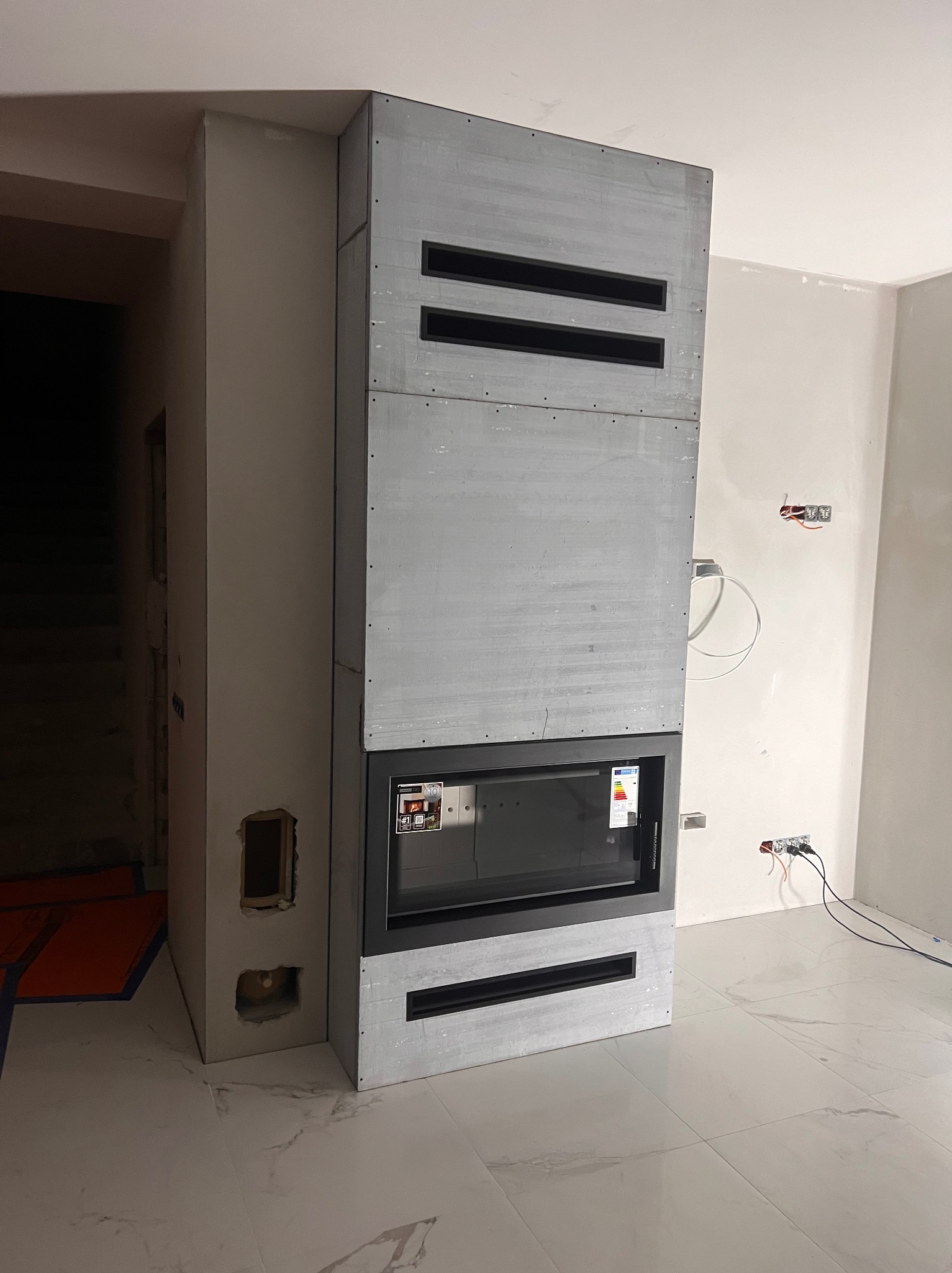Szary, surowy obudowany wkład kominkowy z poziomymi szczelinami wentylacyjnymi, w trakcie montażu, widoczne niedokończone ściany i instalacje elektryczne.