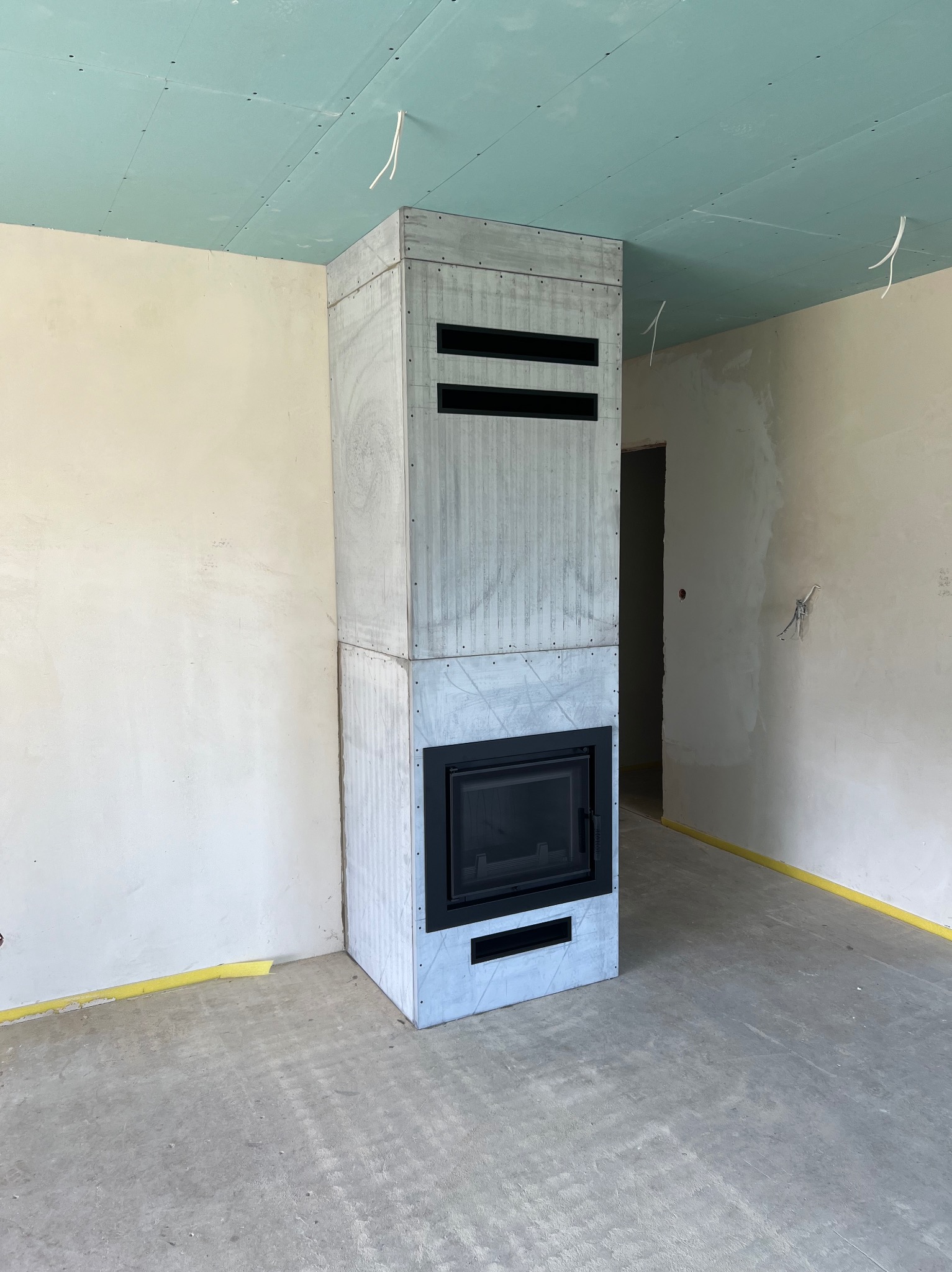 Surowy, betonowy korpus z czarnym wkładem kominkowym w trakcie montażu w pomieszczeniu z surowymi ścianami i sufitem z płyt gipsowo-kartonowych.