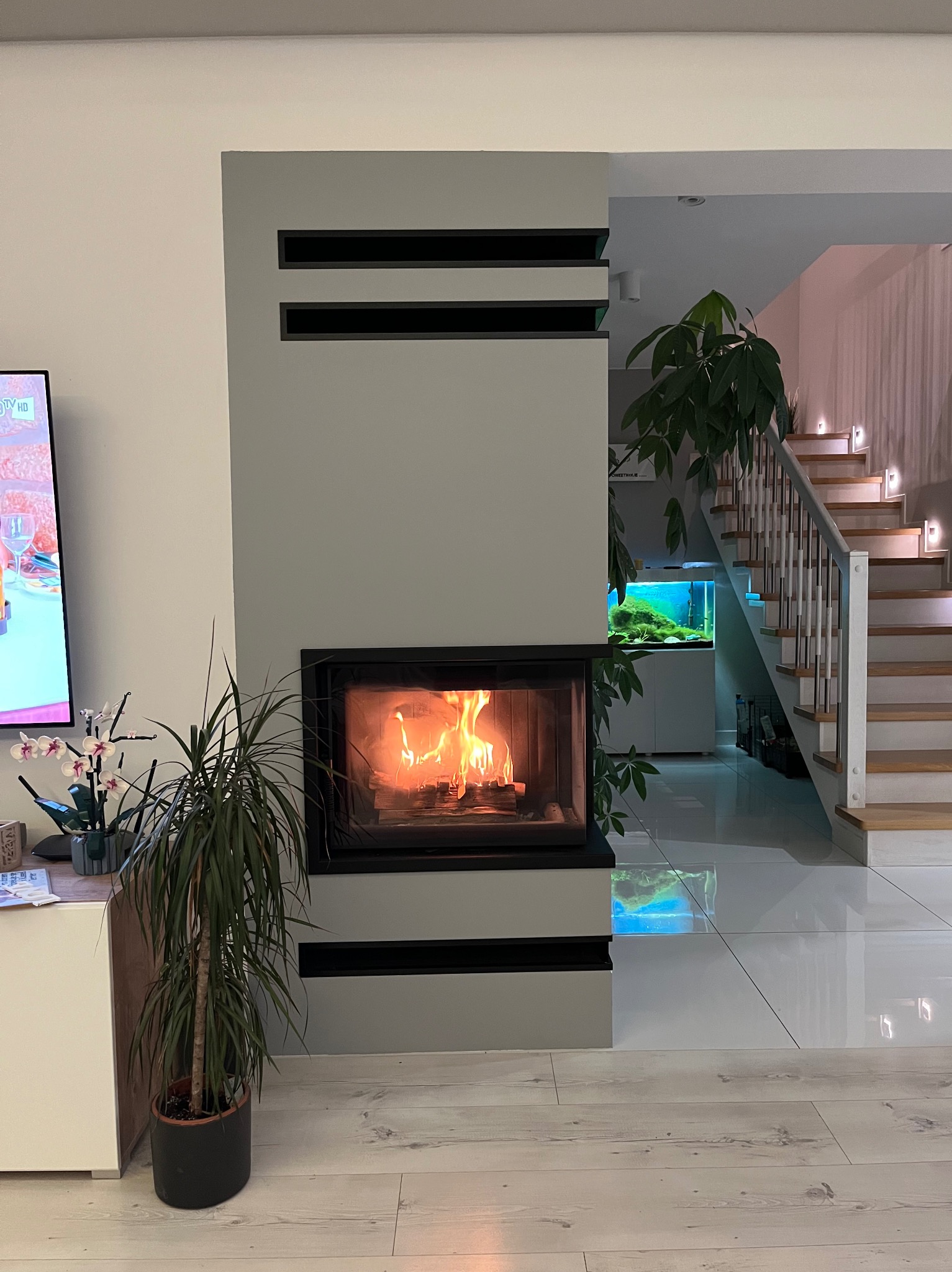 Nowoczesny salon z wbudowanym paleniskiem, widok na schody i akwarium w tle.